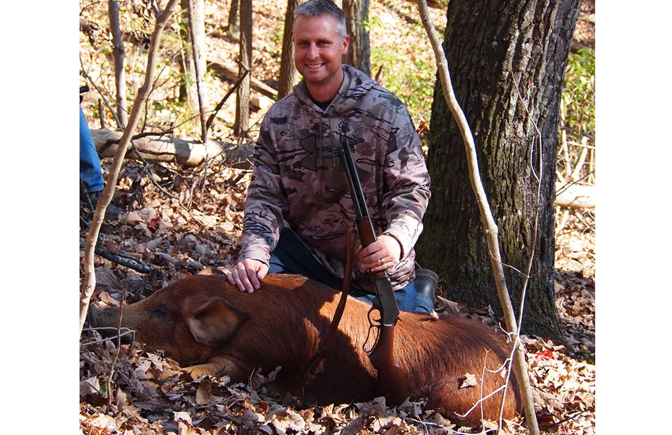 Feral hog on the ground in wooded outdoor setting, person in tactical gear kneeling next to it
