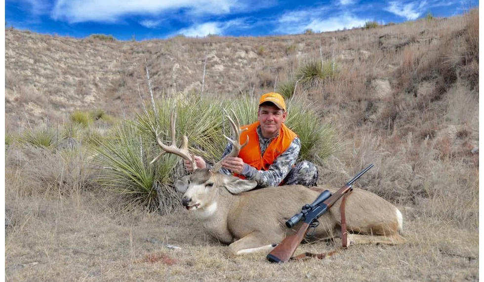 Hunter with Henry repeating rifle posing next to trophy white-tailed deer in rugged outdoor setting