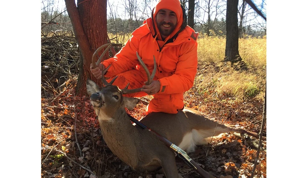 Man in orange jacket and camouflage pants poses with a deer he just hunted using a Henry rifle
