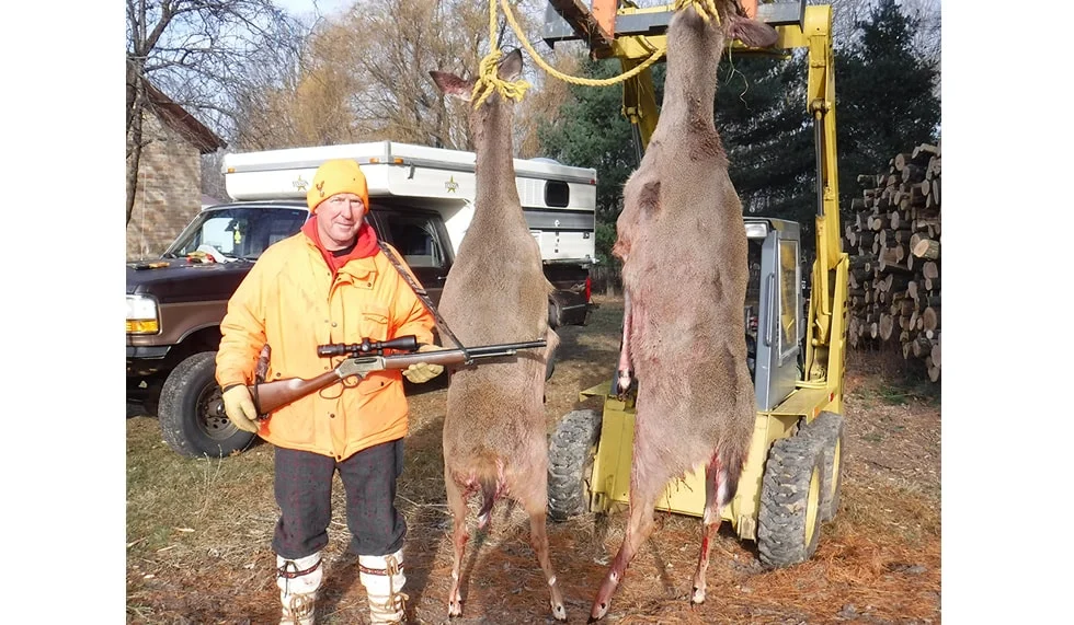 Hunter in yellow jacket holding rifle next to large deer carcass hanging from machinery on rural property
