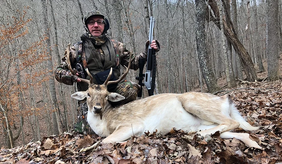 Hunter in camouflage gear holding a rifle and posing with a deer he hunted in a forested area