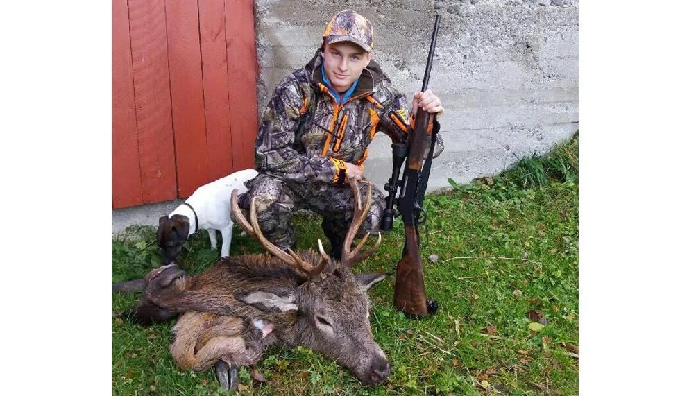 A hunter wearing camouflage gear and holding a rifle poses next to a large white-tailed deer