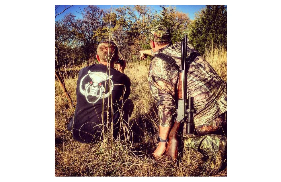 A person carrying a rifle stands in a grassy field with trees and bushes in the background, preparing to hunt a hog