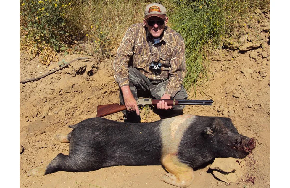 Camouflaged hunter with a rifle standing next to a killed wild boar in a natural outdoor setting