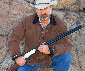 Cowboy with lever-action rifle in outdoor setting, wearing brown jacket and cowboy hat