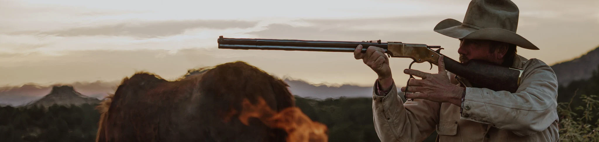 Man aiming a lever-action rifle in a scenic outdoor landscape with mountains in the background