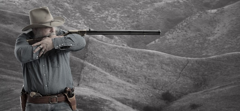 Bearded man aiming a New Original Henry rifle using a cross-armed support stance.