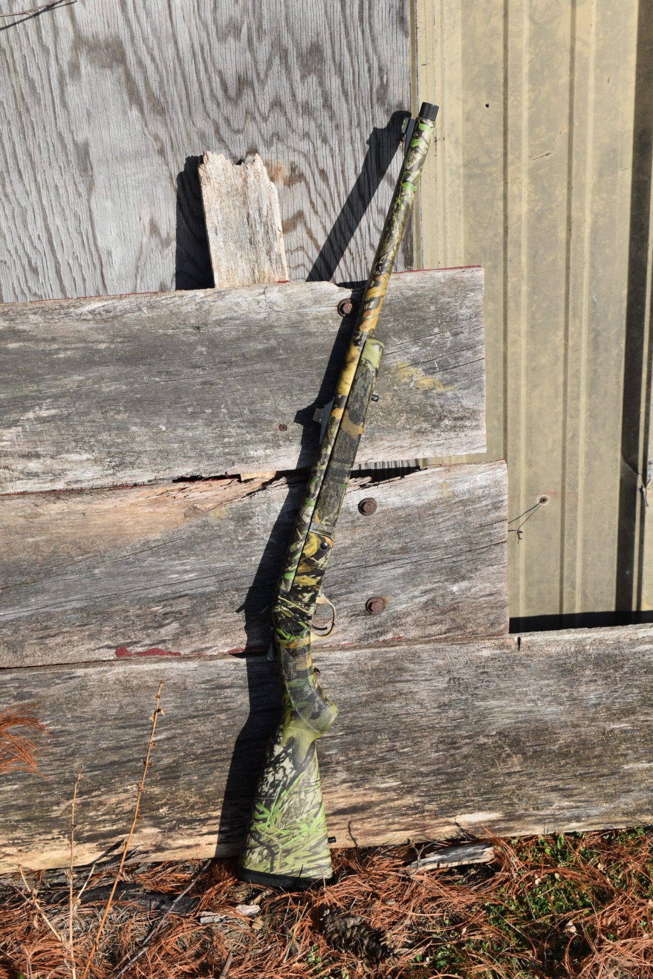 Camouflaged single shot shotgun leaning up against a stack of wood