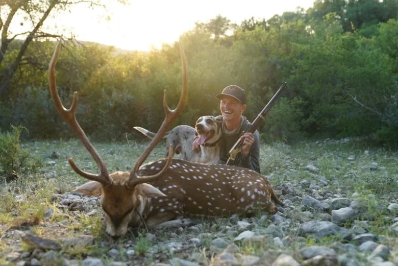 Hunter poses with axis deer in natural outdoor setting at sunset