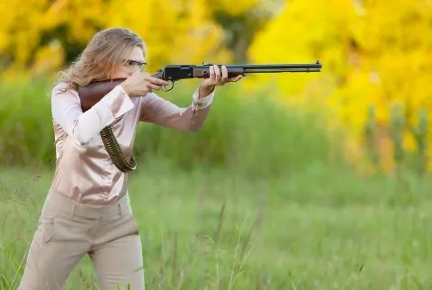 A woman in a pink blouse with a bandoleer aiming a pump action rifle
