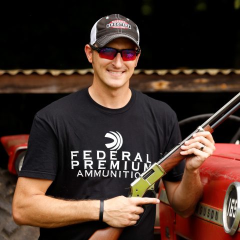 Photo of a man standing next to a vintage tractor smiling and holding a rifle.