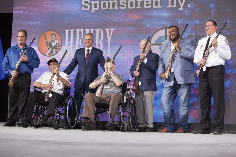 Anthony Imperato standing in the middle of a line of veterans on a stage holding rifles.