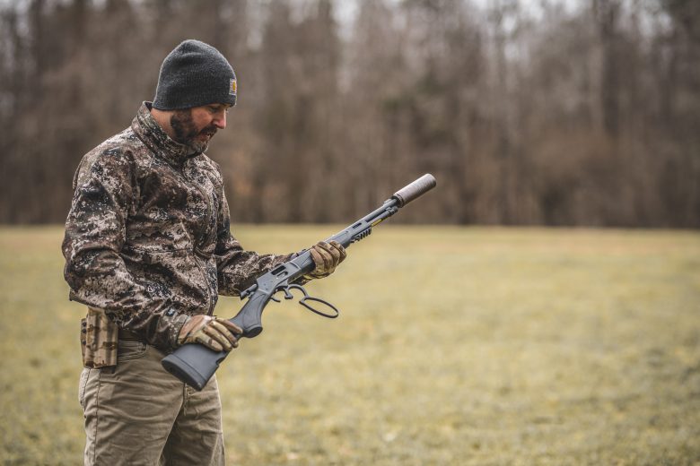 Man dressed in camo clothing holding a rifle with a suppressor on the end of it.