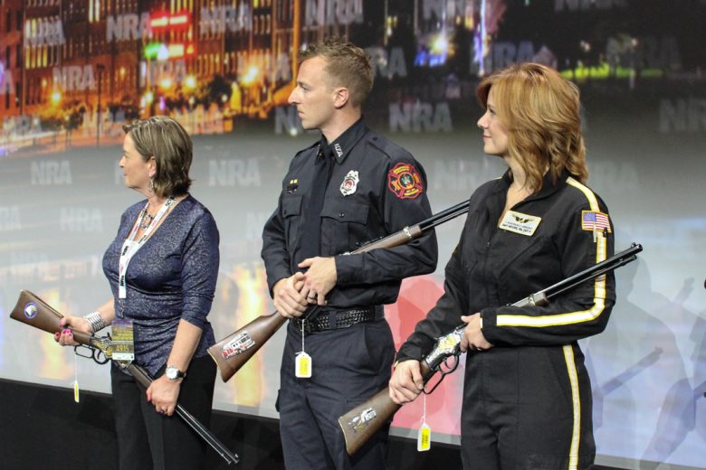 Recipients of a rifle presentation stand on stage holding their rifles and listening to a speech.