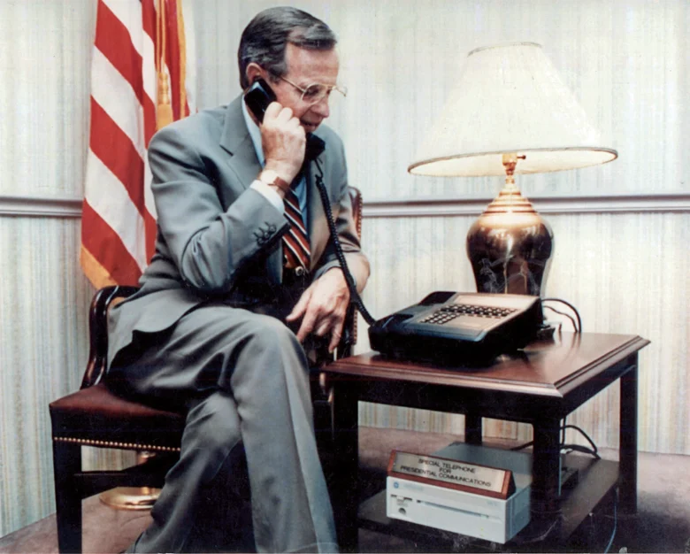 Businessman in a suit speaking on a vintage-style telephone in an office setting, with a US flag in the background