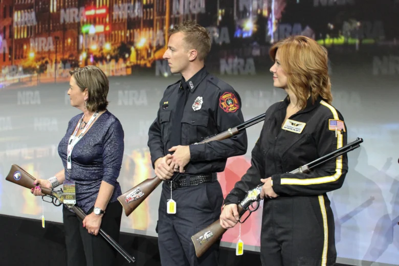 Three people, two women and one man, standing on stage holding Henry lever-action rifles and listening to a speech.