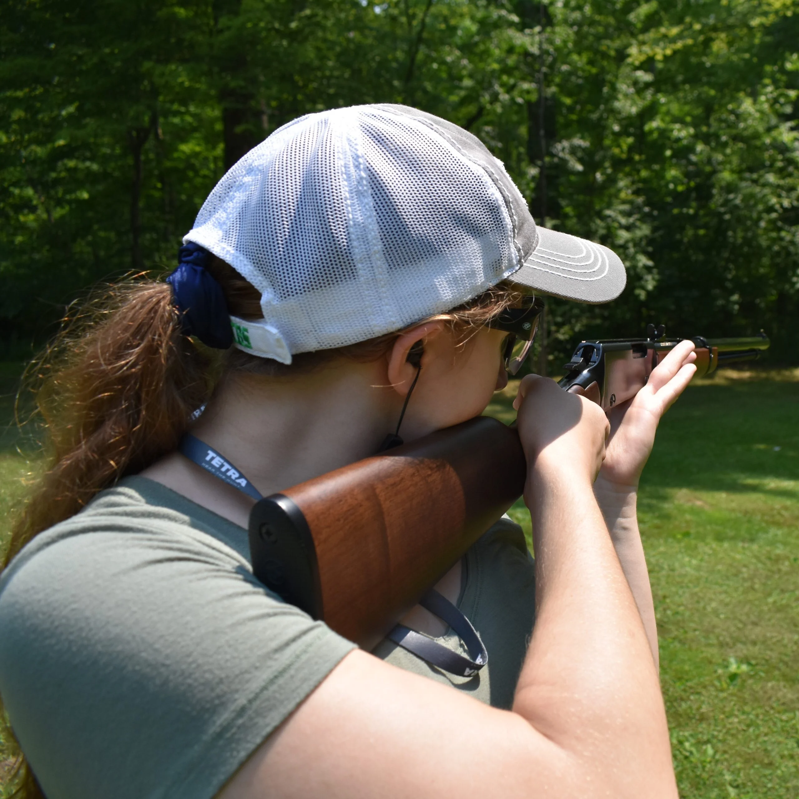 Person wearing a baseball cap and tactical gear shooting a rifle offhand in a wooded outdoor setting
