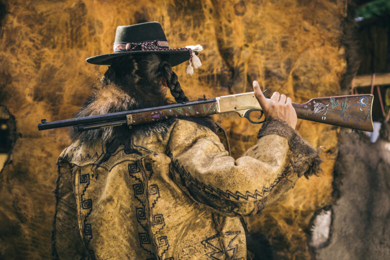 Man in a handmade leather coat holding a rifle over his shoulder looking away from the camera.