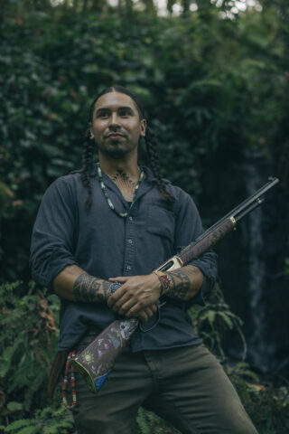 A man smiling holding a painted rifle in his arms with a forest in the background.