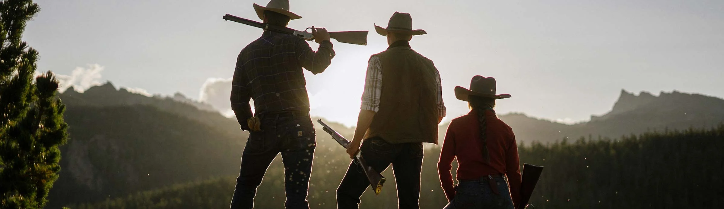 Silhouettes of three cowboys holding Henry rifles against a scenic mountain landscape at sunset