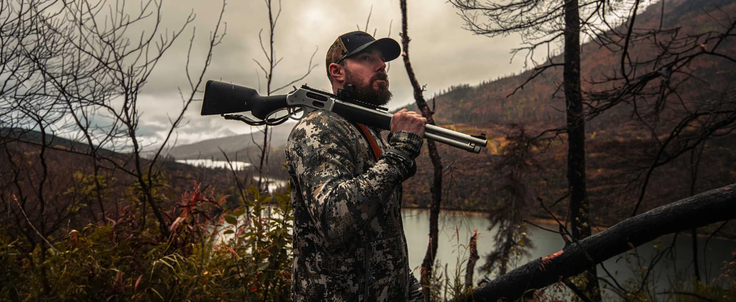 Hunter in digital camouflage with a double-barrel shotgun slung over his shoulder, standing in a leafless forest by a lake.