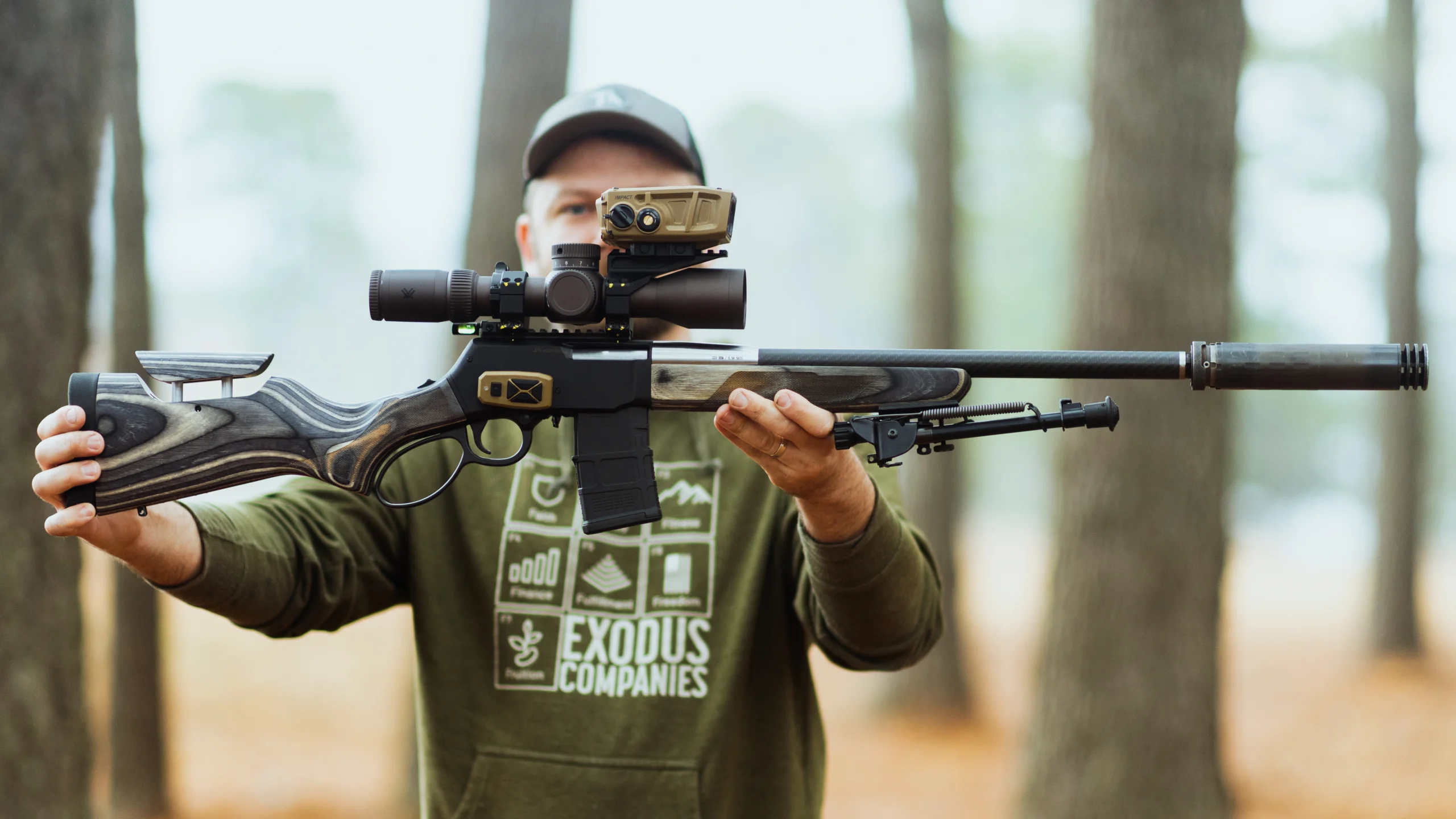 Person holding a rifle with scope aimed forward in a forest setting, wearing a cap and green hoodie.