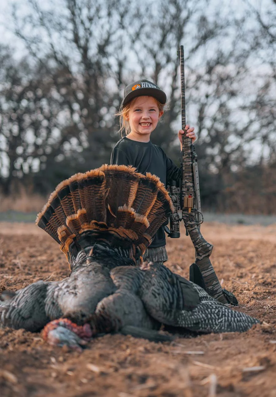Young girl hunter posing with a camo-cap and rifle beside a large dead turkey on a dirt field.