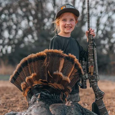 Smiling young girl outdoors wearing a cap, holding a camouflaged rifle beside a large turkey tail fan.