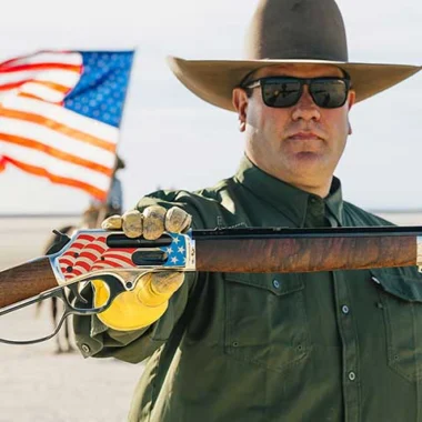 Man wearing a wide-brim hat and sunglasses holds a rifle with an American flag design on the stock, outdoors with a flag in the background.