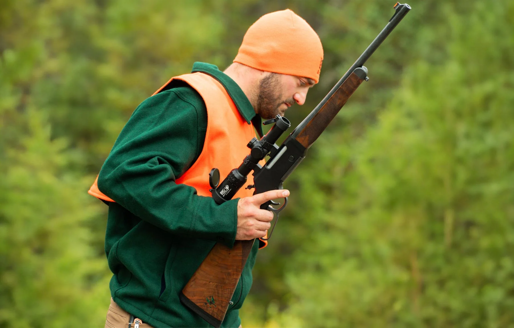 Man wearing an orange safety vest and green jacket aims a scoped rifle outdoors with a forest background.