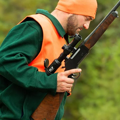 Hunter wearing an orange safety vest and cap, examining a scoped rifle in a forested area.