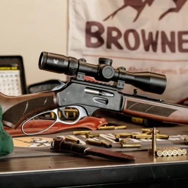 A scoped rifle resting on a table with spent cartridges and a green pouch nearby.
