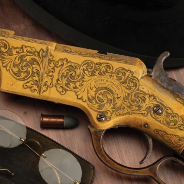 Ornate gold-colored pistol with intricate scrollwork engraving, a single bullet nearby on a wooden surface.