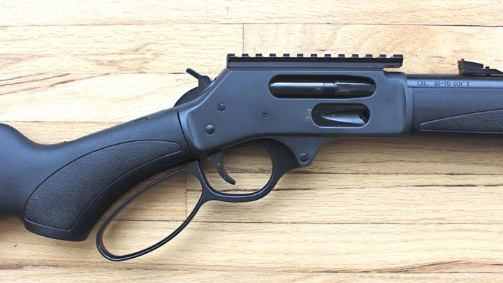 Close-up of a black lever-action rifle lying on a light wood surface, showing the trigger guard and receiver.