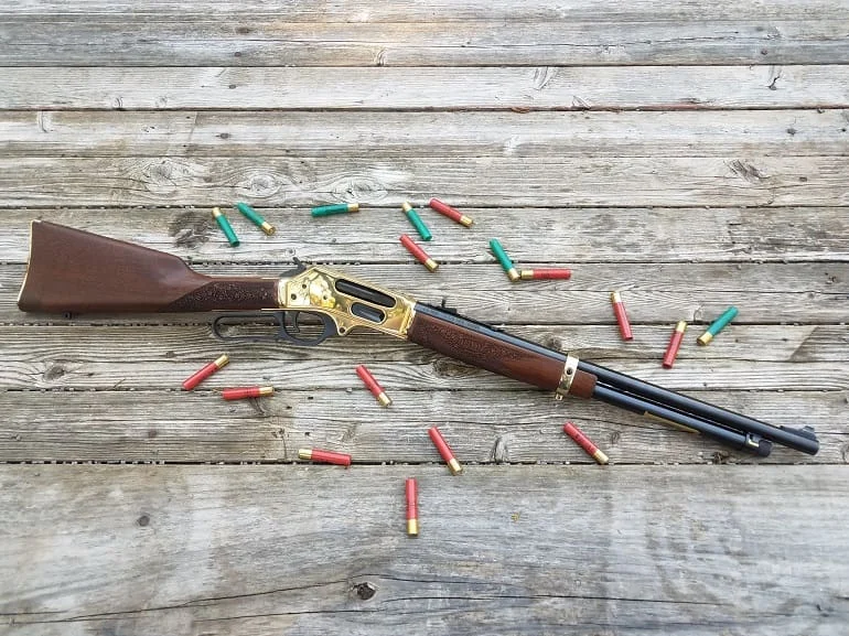 Double-barrel shotgun with wooden stock and brass receiver lies on a weathered wooden surface, with red and green spent shell casings scattered around.