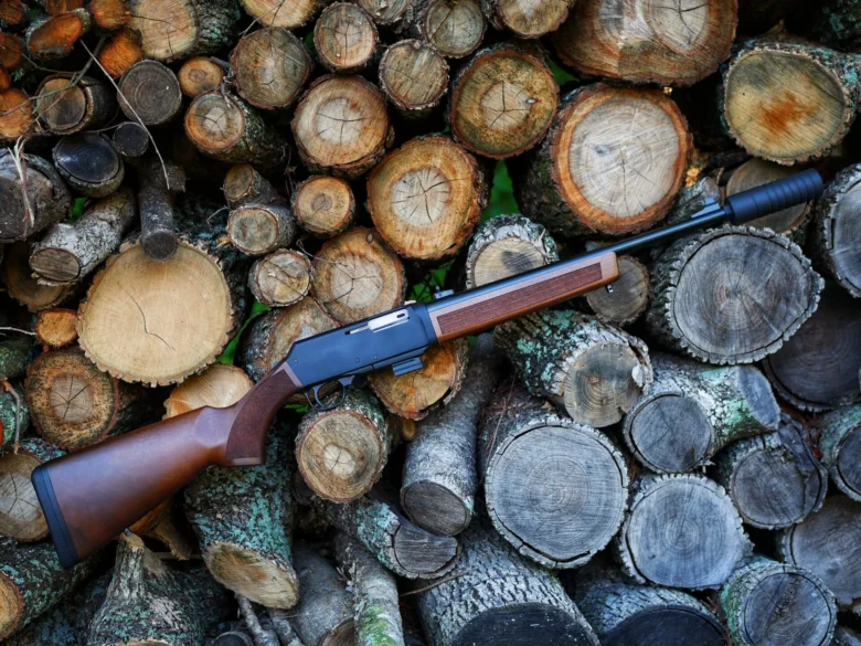 Rifle lying across a stack of cut logs in a wooded area, with a wooden stock and dark barrel visible