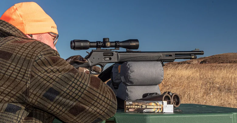 Shooter in an orange beanie and plaid jacket aims a scoped rifle resting on a front bag at an outdoor shooting range.