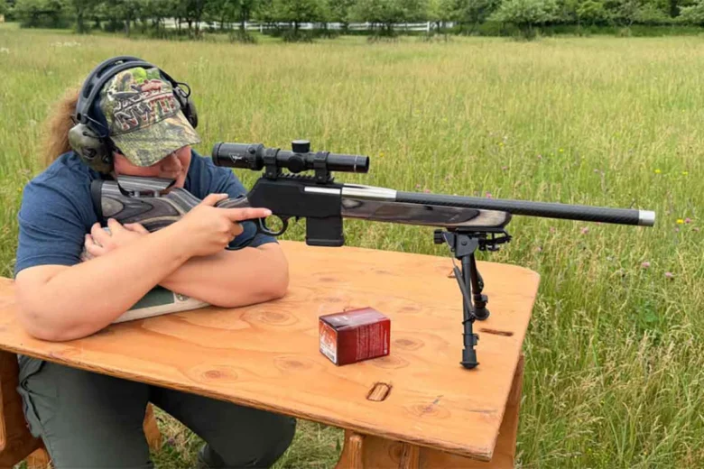 Adult shooter wearing a camo cap and hearing protection rests a scoped bolt‑action rifle on a wooden bench at an outdoor firing range.