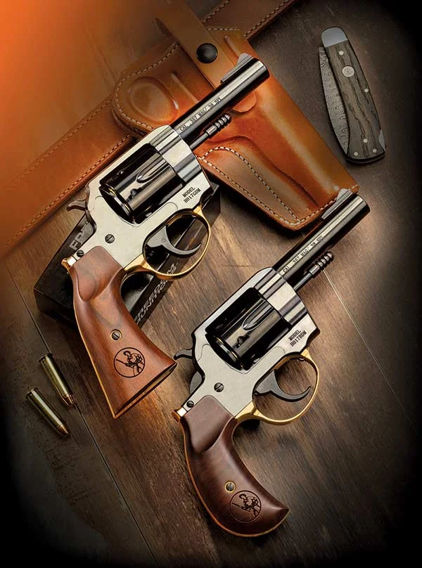 Two revolvers with wooden grips and silver frames lie on a dark wooden surface beside a brown leather holster and a small folding knife.