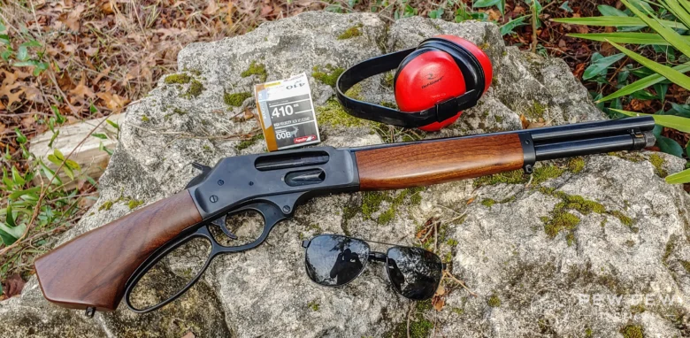 Lever-action rifle with wooden stock lying on a mossy rock, beside a box of .410 ammo and red earmuffs.