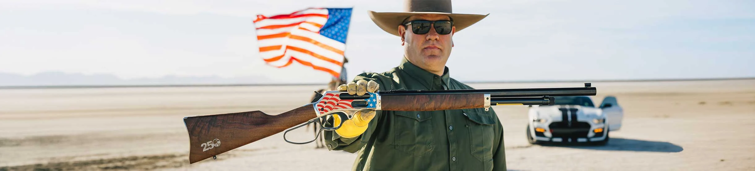 Man in sunglasses and a wide-brimmed hat presents a lever-action rifle decorated with an American flag motif in a desert setting, with a white car in the background.