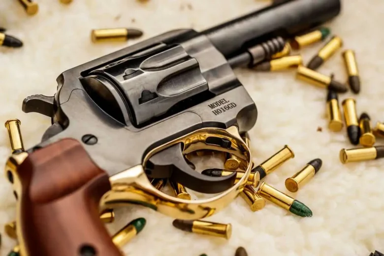 Gold ring wrapped around a handgun, surrounded by scattered bullets on a light surface.