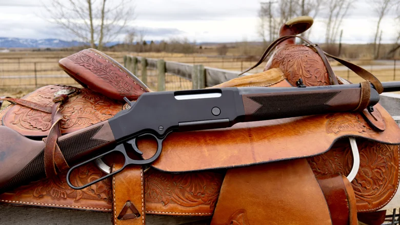 Lever-action rifle resting on a carved leather saddle outdoors, with fence and fields in the background.