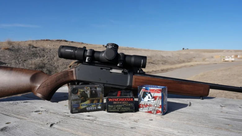 Bolt-action rifle with a scope resting on a weathered wooden bench, three ammo boxes in front, outdoor shooting range in the background.