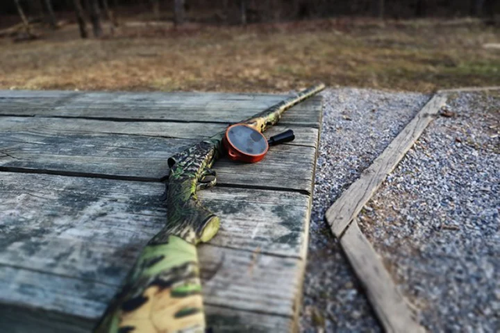 Camouflage hunting rifle with a red-scope resting on a weathered wooden table outdoors.