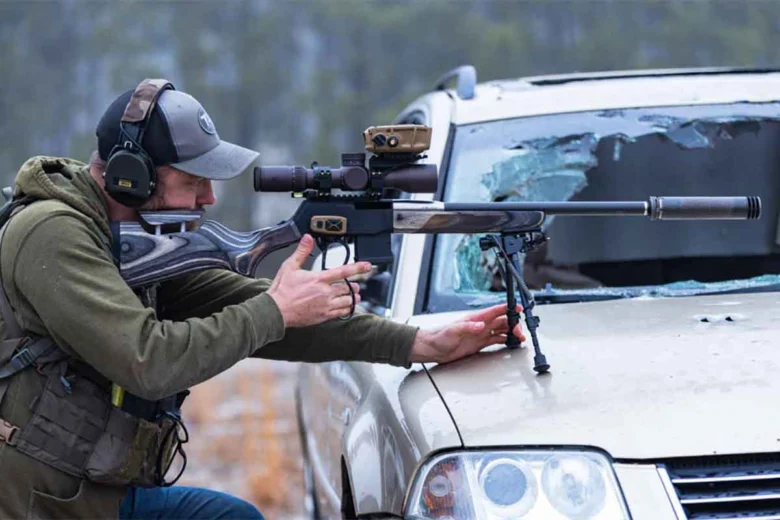 Man wearing a cap, ear protection, and tactical vest aims a scoped rifle resting on the hood of a damaged SUV, with a shattered windshield visible in the background.