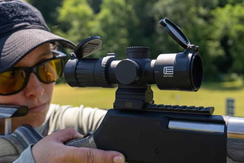 Person wearing a cap and sunglasses aiming a rifle with a scope outdoors at a range/field nearby.