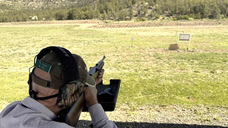 Person at an outdoor shooting range aiming a rifle, wearing ear protection, hat, and a face mask with a camo pattern; targets visible in the field ahead.
