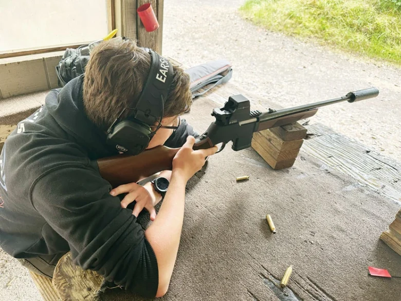 Person at an outdoor shooting range aiming a rifle, wearing hearing protection and a watch, resting on a wooden bench.
