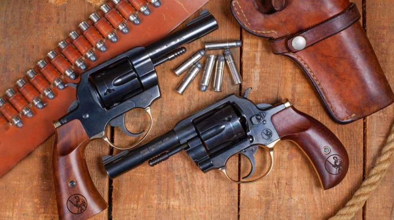 Two revolvers with wooden grips on a wooden table, plus scattered bullets and a leather holster nearby.